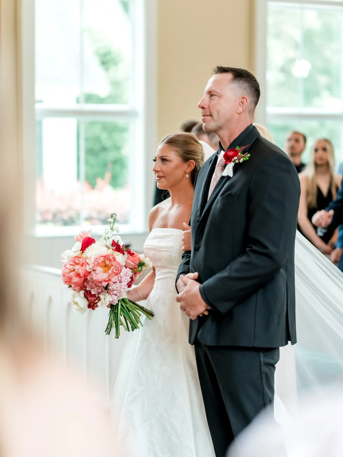 Bride and groom at the altar of a light-filled chapel, the bride holding a coral peony and red ranunculus bouquet