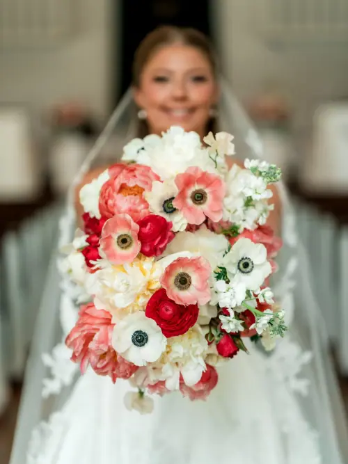 Coral peony, white anemone and red ranunculus bridal bouquet held toward the camera in a chapel aisle