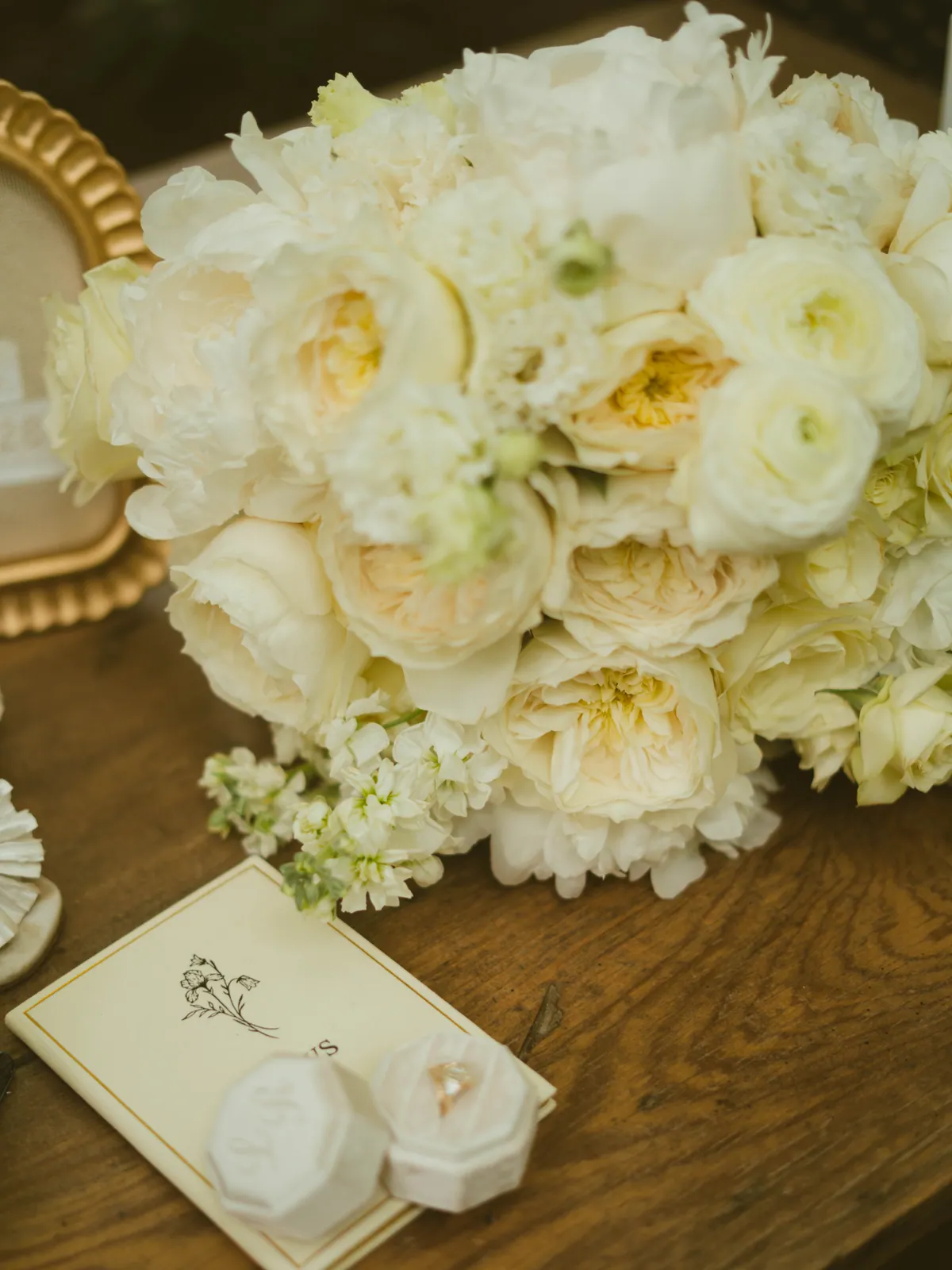 Ivory garden rose and ranunculus bridal bouquet resting on a wooden table beside the vow book and two ring boxes