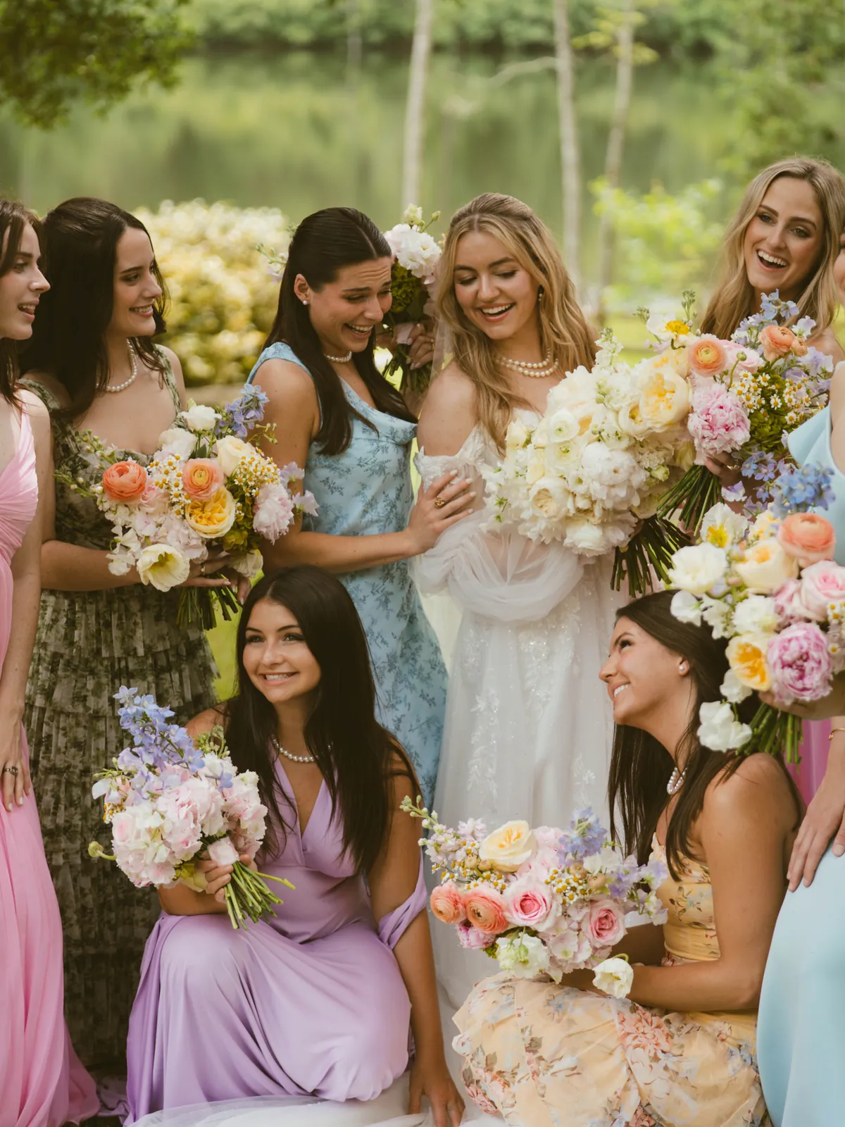 Bridesmaids in pastel gowns holding bouquets of peach ranunculus, lilac stock and white garden roses beside a lake