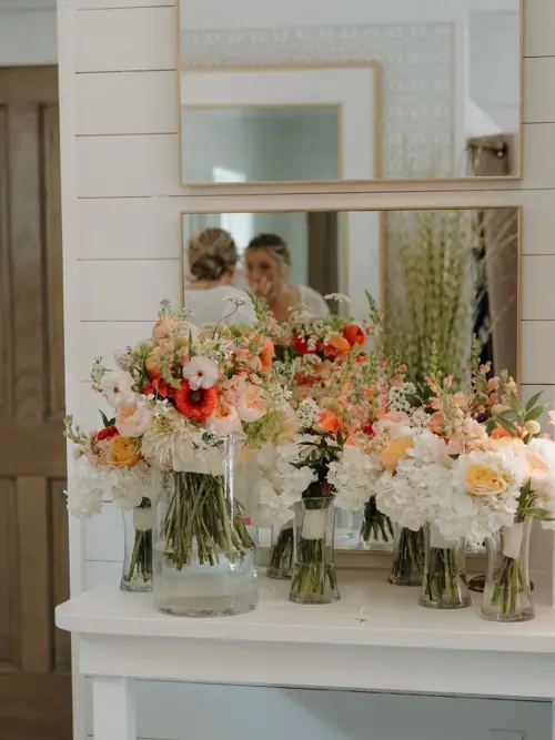 Six bridal party bouquets in glass vases lined along a getting-ready vanity in peach, coral and cream