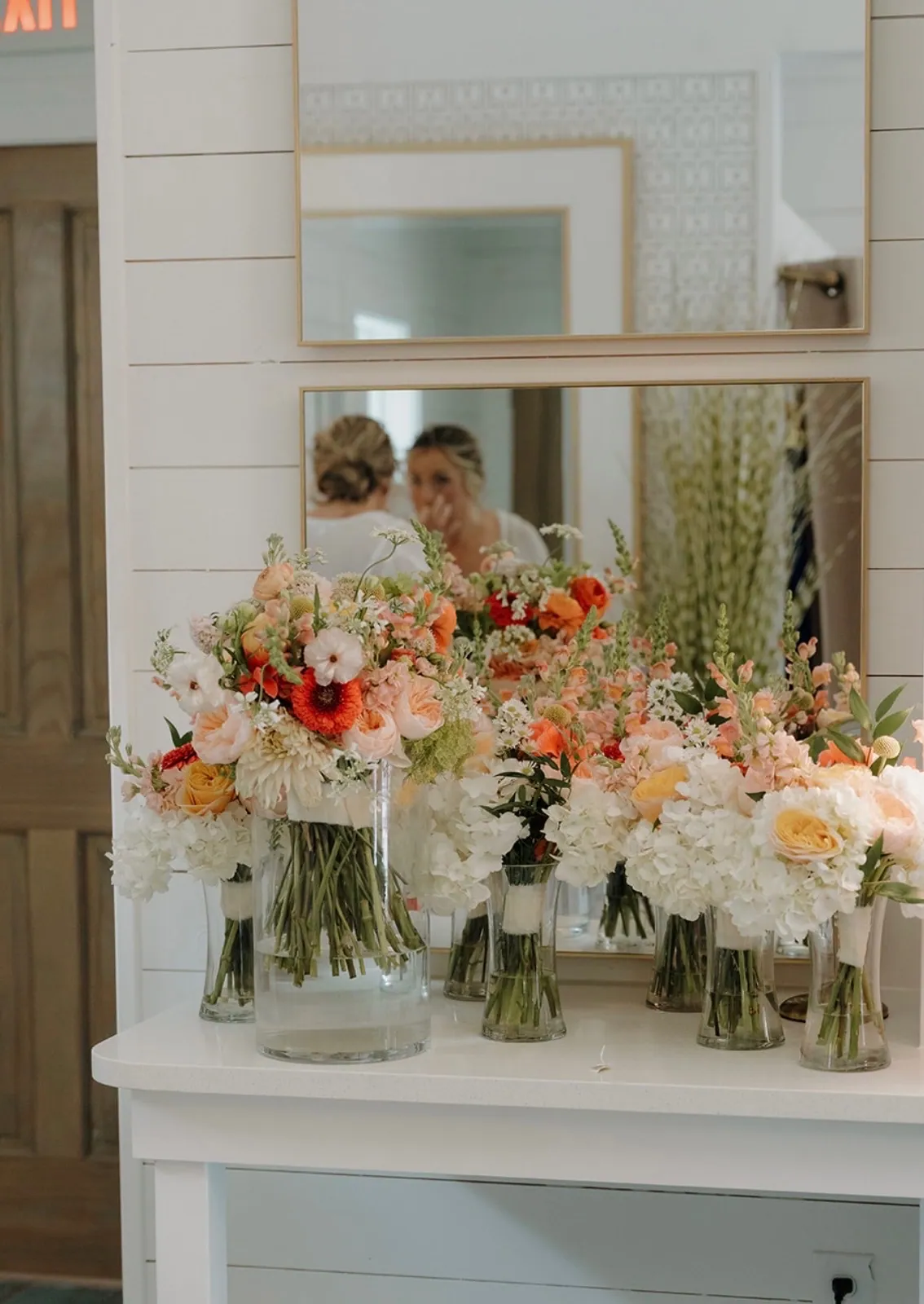 Six bridal party bouquets in glass vases lined along a getting-ready vanity in peach, coral and cream