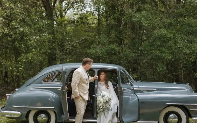 Bride carrying a white and green bouquet stepping out of a vintage car under live oaks