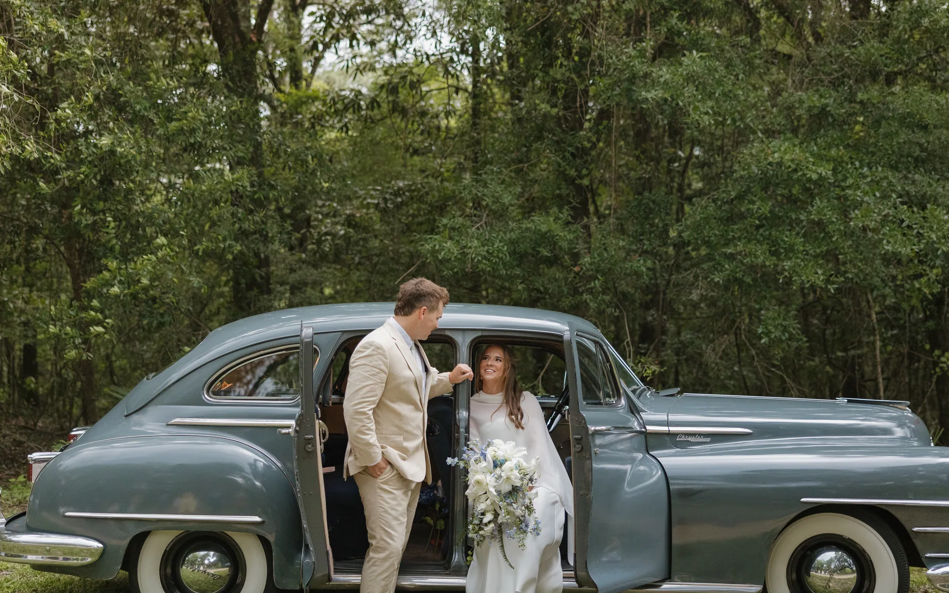 Bride carrying a white and green bouquet stepping out of a vintage car under live oaks