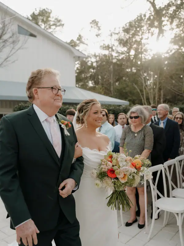 Father walking his daughter down an outdoor aisle, the bride carrying a loose peach and coral bouquet