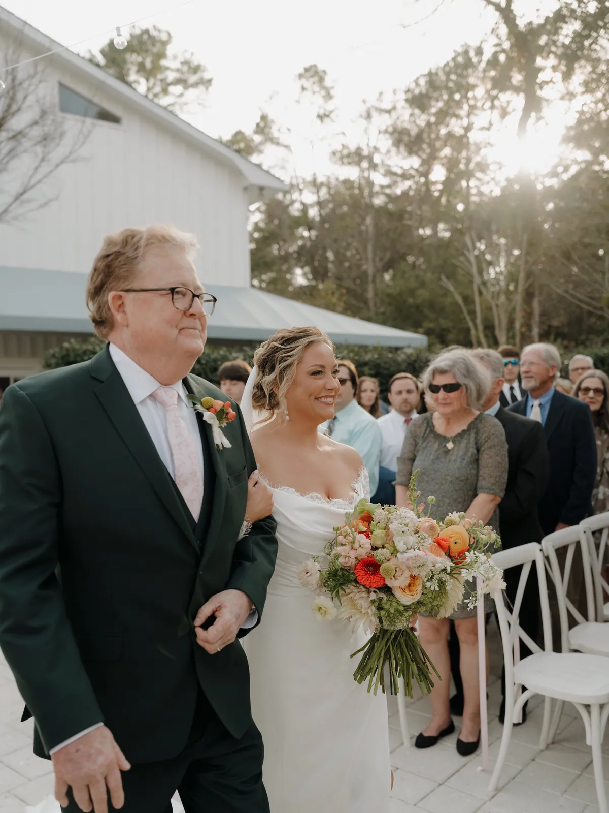 Father walking his daughter down an outdoor aisle, the bride carrying a loose peach and coral bouquet
