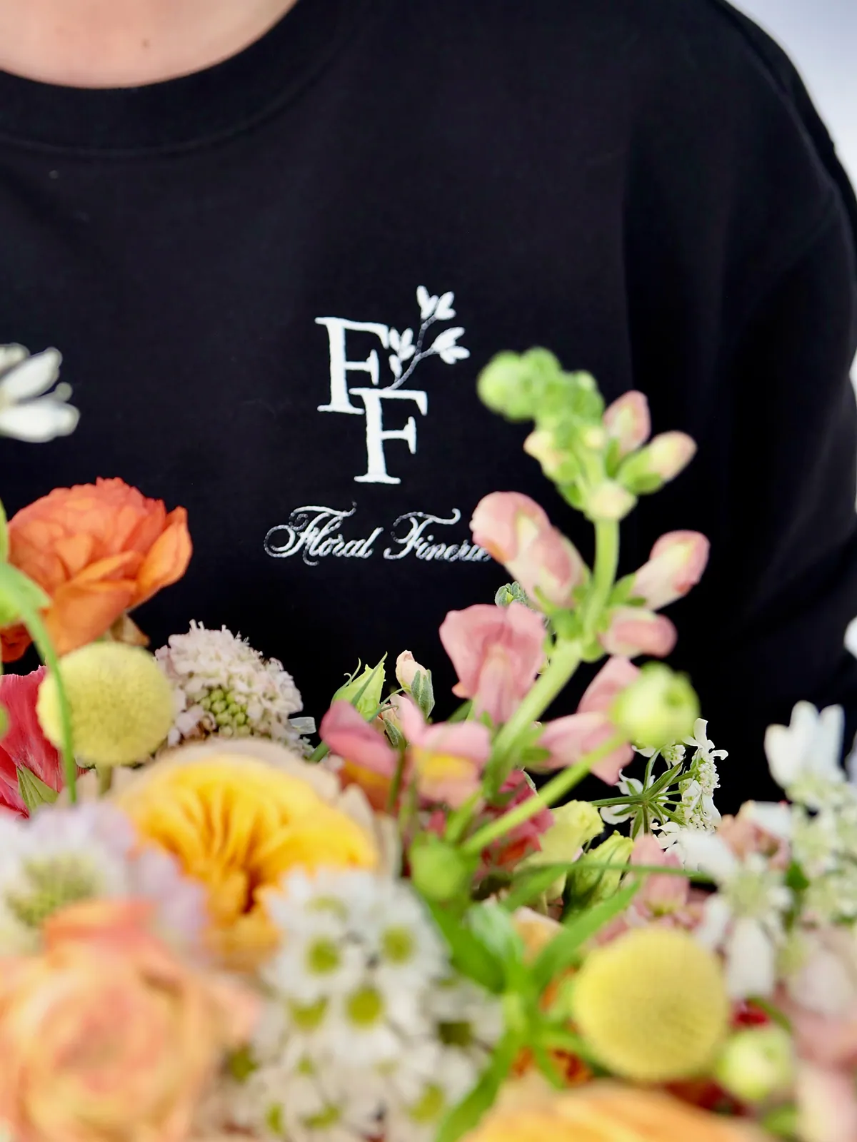 Florist in a Floral Finerie sweatshirt holding a wide arrangement of orange ranunculus, snapdragon and craspedia