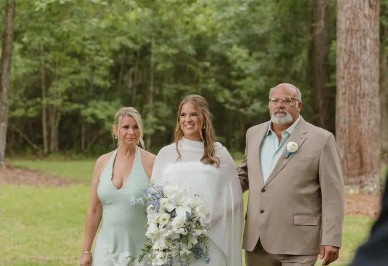 Bride walking between her parents through pine woods, carrying a loose white and blue bouquet