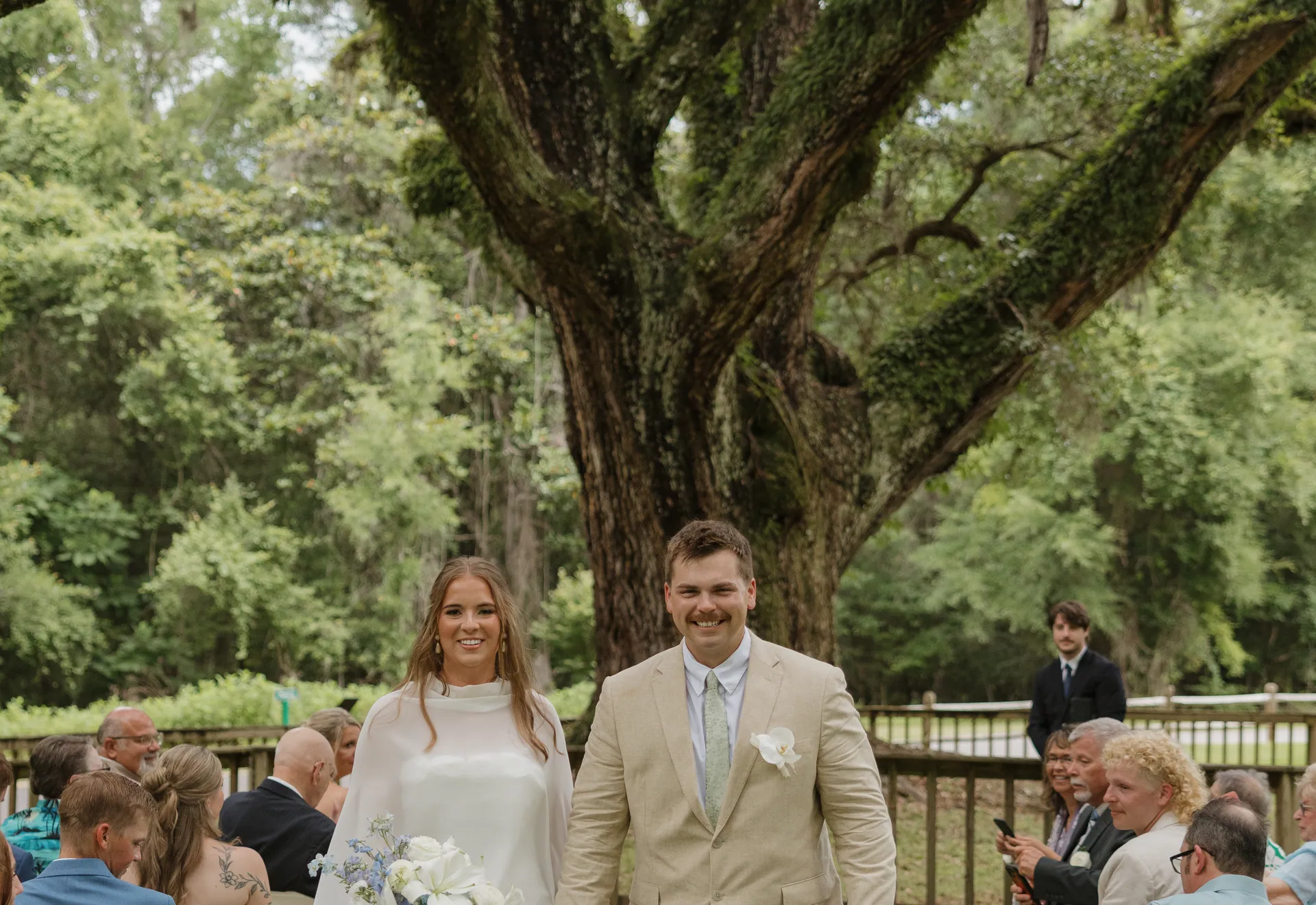Newly married couple walking back up the aisle under a moss-draped live oak, the bride carrying a white and blue bouquet
