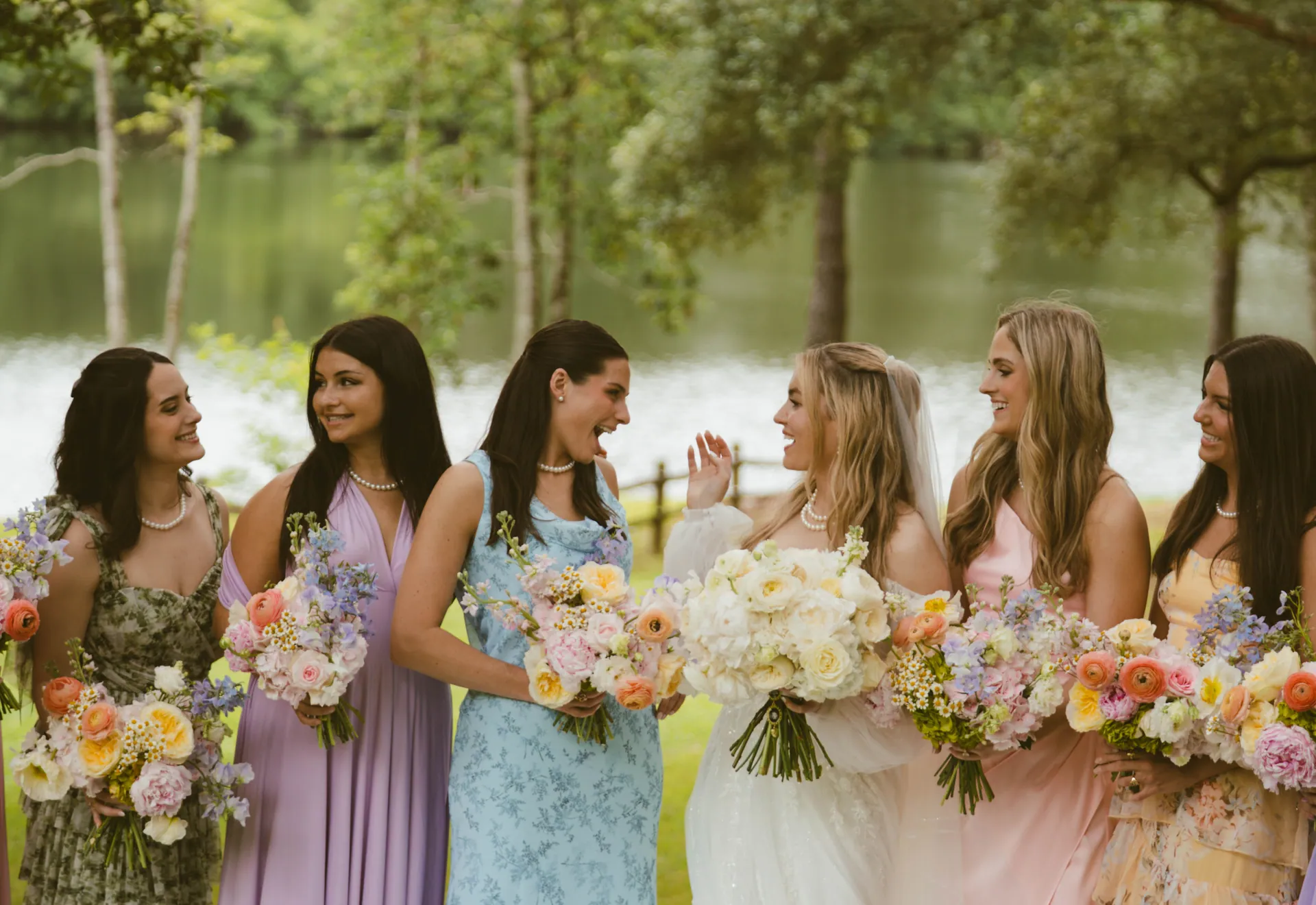 Bride and six bridesmaids by a lake, each carrying a pastel bouquet of ranunculus, sweet pea and garden roses