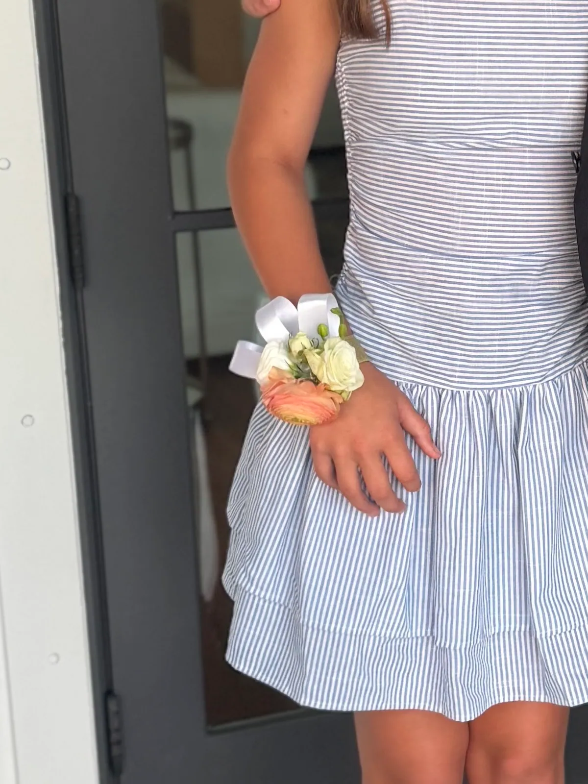 Peach and cream rose wrist corsage on a white ribbon, sized for a child's wrist, worn over a blue striped dress