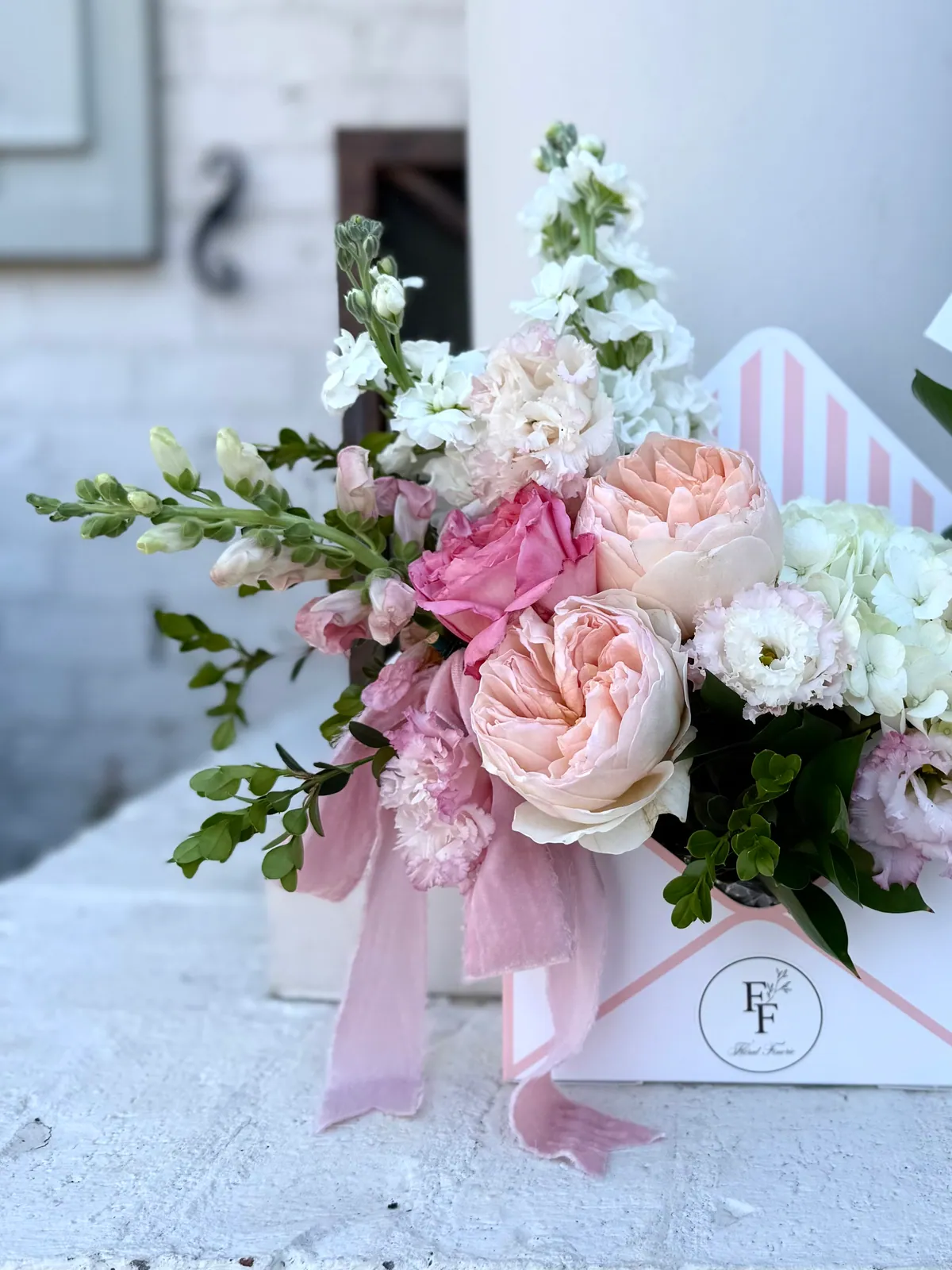 Floral Finerie envelope box of blush garden roses, white stock and snapdragon finished with a pink chiffon ribbon