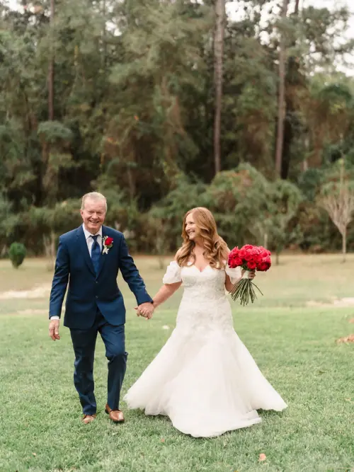 Bride walking across a lawn hand in hand with her father, carrying a full red rose bouquet