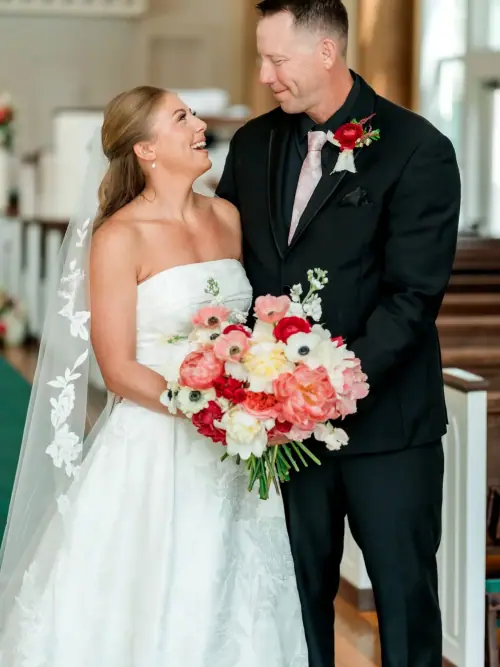 Bride and groom laughing in a church aisle, her bouquet of coral peonies, white anemones and red ranunculus between them