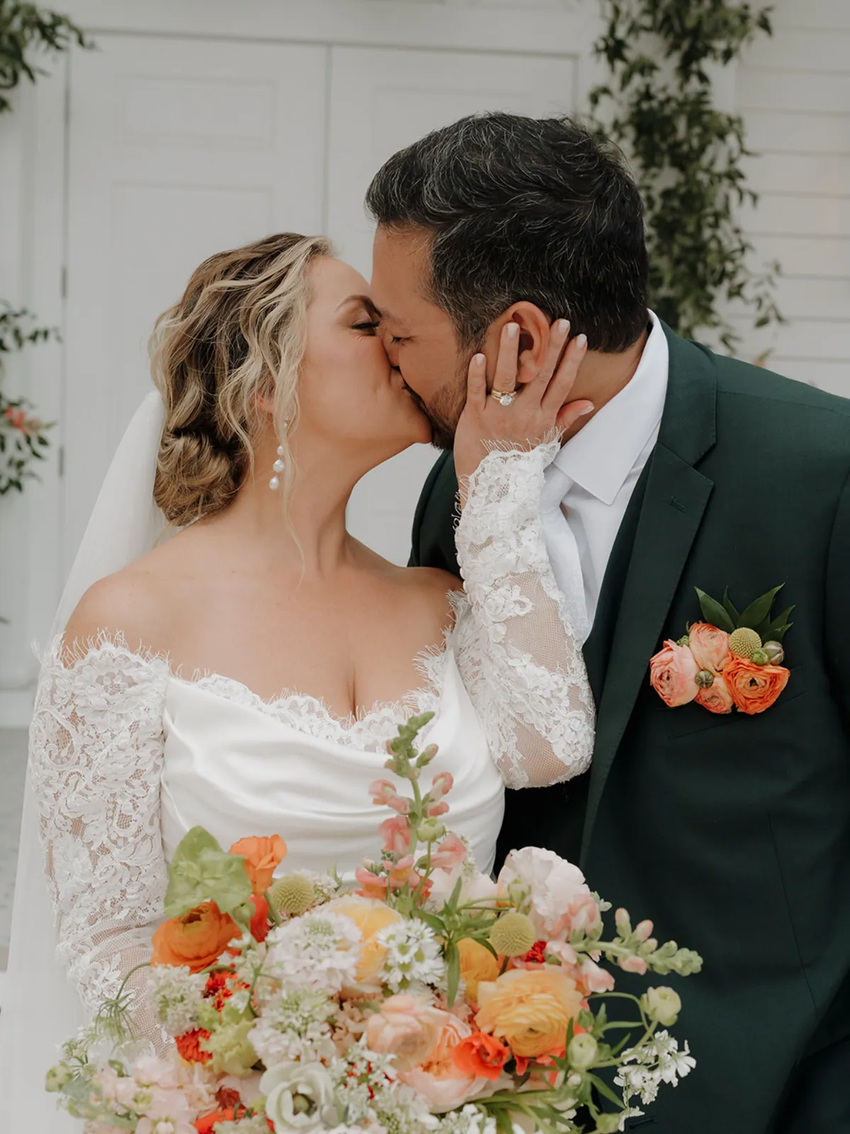 Bride and groom kissing, her peach and orange garden rose bouquet and his matching ranunculus boutonniere in frame