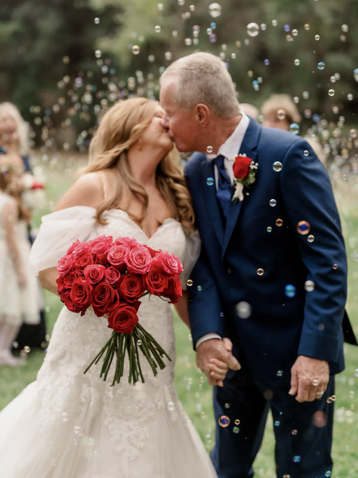 Bride carrying a tightly gathered red rose bouquet with long bound stems as bubbles are blown around the couple