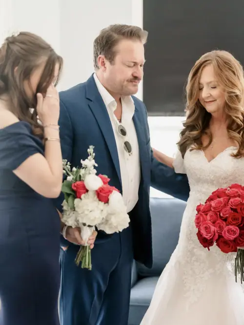 Bride holding a deep red rose bouquet beside a guest carrying white stock and red roses