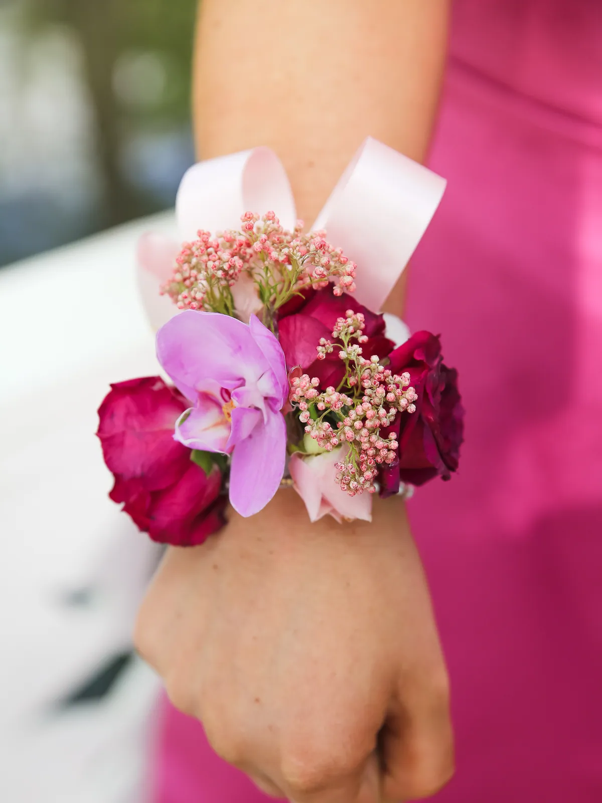 Wrist corsage of magenta roses, a lavender orchid and pink waxflower on a blush ribbon against a fuchsia gown
