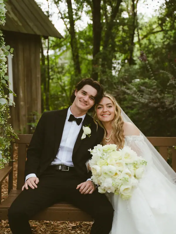 Bride and groom seated on a wooden bench, the bride holding a large all-white bouquet of garden roses and hydrangea