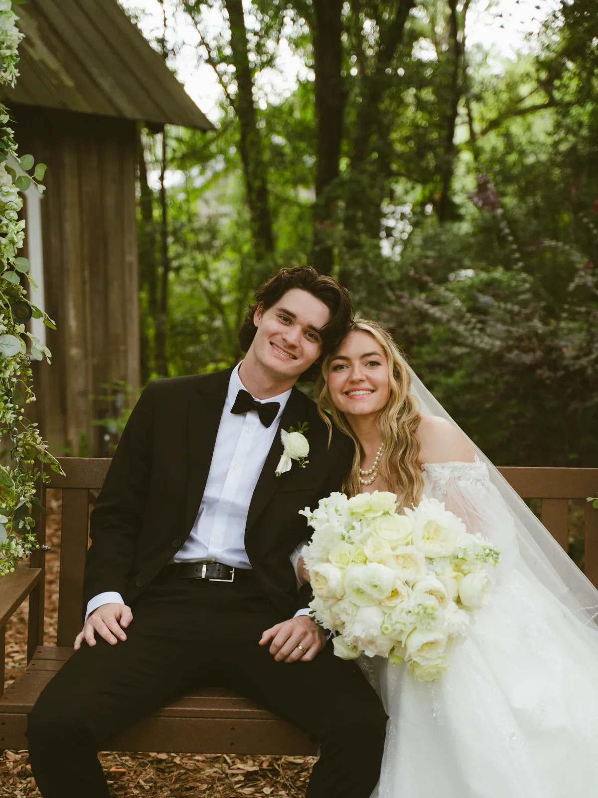 Bride and groom seated on a wooden bench, the bride holding a large all-white bouquet of garden roses and hydrangea